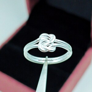 May include: A silver ring with a knot design. The ring has a double band and a knot-shaped centrepiece. The ring is displayed against a black background inside a pink box. The text "SilverGoldWorks" is visible on the ring.