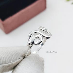 Könnte beinhalten: Ein Ring aus poliertem Silber mit einem gewundenen Schlangendesign, das sich um den Finger legt. Die Schlange hat eine glatte, reflektierende Oberfläche mit einem deutlich erkennbaren Kopf und Schwanz. Der Text SilverGoldWorks ist auf dem Ringband sichtbar.
