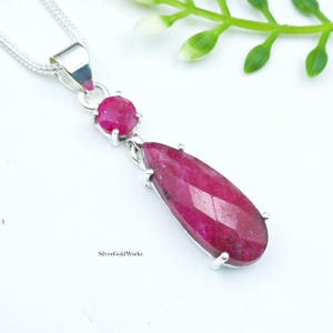 Può includere: Una collana d'argento con un pendente con una grande gemma di rubino a forma di goccia e un rubino rotondo più piccolo. Il pendente è appeso a una delicata catena d'argento. Il rubino è di un colore rosso intenso e ricco.