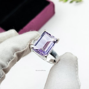 May include: A silver ring featuring a large, rectangular, faceted light purple gemstone held by four prongs. The band is smooth and polished, showcasing a clean design. The text SilverGoldWorks is printed in small black letters near the base.