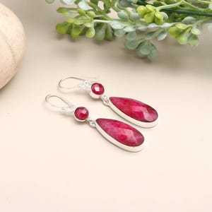May include: Silver earrings with a teardrop design. Each earring has a small, round, red gemstone at the top and a larger, faceted, red gemstone in a teardrop shape. The earrings are displayed on a light-coloured surface.