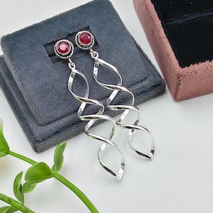 May include: A pair of long silver earrings featuring a spiral, twisted metal design. Each earring is topped with a round, faceted dark red gemstone set in a twisted silver frame. The earrings have a polished, reflective finish and a dangling structure.
