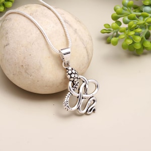 Puede incluir: Collar con colgante plateado con diseño de serpiente. El colgante tiene cabeza y cuerpo texturizados, con intrincados patrones de bucles. El collar se muestra sobre una piedra de color claro, con una cadena sencilla.