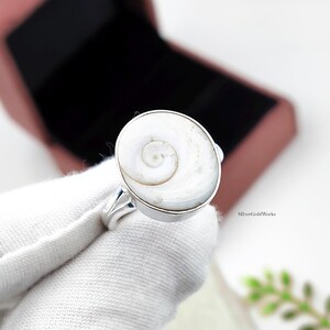 Könnte beinhalten: Ein Silberring mit einer runden Einlage aus weißem Shiva-Auge-Muschelgehäuse, das ein natürliches Spiralmuster aufweist. Die Muschel ist in eine polierte Silberfassung mit einem schlichten Ringband eingefasst. Der Text "SIlverGoldWorks" ist sichtbar.