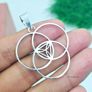 May include: A silver pendant featuring a geometric design of overlapping circles and a central triangular pattern. The pendant has a loop for a chain and is held in a hand. The pendant is a piece of jewelry.