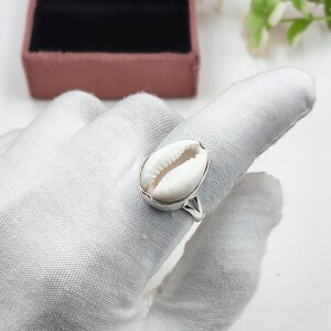 Peut inclure: Une bague en argent ornée d'un coquillage cauri blanc naturel en son centre. Le coquillage présente une forme ovale lisse avec une fente centrale, monté solidement sur une monture en argent poli qui entoure le doigt avec un anneau simple.