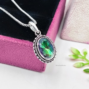 Puede incluir: Un collar con colgante de plata que presenta una piedra preciosa ovalada e iridiscente engastada en un marco de plata con diseño de cuerda trenzada. El colgante cuelga de una fina cadena de plata. El texto SilverGoldWorks aparece en letras negras.