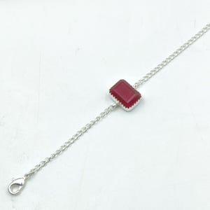 Pode incluir: Uma pulseira de corrente de prata com uma única pedra preciosa retangular facetada de cor vermelho-escura, engastada numa moldura de prata serrilhada. A pulseira inclui um fecho lagosta numa extremidade que liga os elos metálicos.