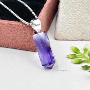 May include: A pendant necklace featuring a faceted, hexagonal purple amethyst crystal point suspended from a silver-toned metal bail and a thin silver chain. The crystal displays natural color variations from deep violet to translucent white.