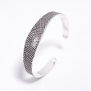 May include: A silver-colored cuff bracelet with a textured, dotted design on the top surface. The bracelet has a smooth, polished finish on the inner and outer edges, and is open at the ends.