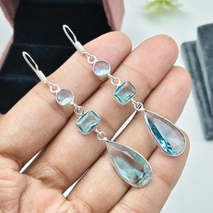 Puede incluir: Un par de pendientes largos con ganchos de plata y una disposición vertical de tres piedras preciosas de color azul claro. Cada pendiente incluye una pequeña piedra redonda, una piedra rectangular y una gran piedra facetada en forma de pera en la parte inferior.