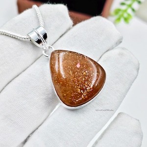 Puede incluir: Un colgante de piedra de sol en forma de lágrima con un tono marrón cobrizo cálido y partículas metálicas brillantes incrustadas. La gema está engastada en un metal de tono plateado y cuelga de una cadena de tono plateado a juego.