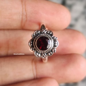 以下が含まれることがあります： 中央に丸い深紅色の宝石をあしらったシルバーリング。宝石は、詳細な花をモチーフにしたセッティングで囲まれています。リングのデザインには、複雑な模様と、ヴィンテージまたはアンティークスタイルを思わせるテクスチャード加工されたバンドが含まれています。