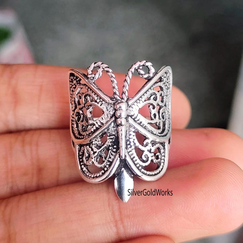 Puede incluir: Un anillo plateado con forma de mariposa con intrincados detalles de filigrana. El anillo presenta un dise&ntilde;o detallado de mariposa con alas caladas y un cuerpo texturizado. El texto "SilverGoldWorks" es visible en el anillo.