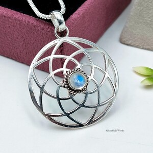 May include: A circular silver pendant featuring an intricate openwork geometric pattern of intersecting arcs. At the center, a small, round, iridescent moonstone is set within a twisted silver rope border. The pendant hangs from a silver chain.