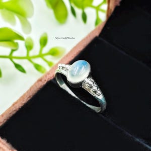 May include: A silver ring with an oval, light blue gemstone. The ring has a detailed band with small decorative elements. The ring is displayed in a dark jewelry box, with green foliage in the background. The text "SilverGoldWorks" is visible.