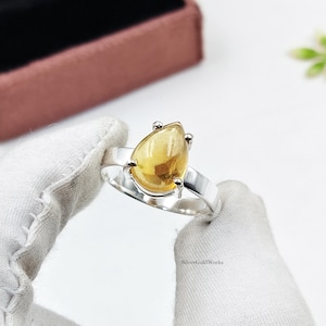 May include: A silver ring featuring a pear-shaped, translucent yellow citrine gemstone held by four prongs. The band is smooth and polished silver. The text SilverGoldWorks is visible on the band. The ring is held by white fabric gloves.