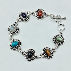 Puede incluir: Una pulsera de eslabones de plata con siete piedras preciosas ovaladas en cabujón de varios colores, incluyendo morado, naranja, blanco, turquesa, negro, marrón y verde. Cada piedra está engastada en un marco de plata con cuentas y un cierre de barra.