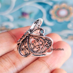 Puede incluir: Primer plano de un anillo plateado con un dise&ntilde;o de mariposa calada. El anillo presenta intrincados detalles de filigrana en las alas y el cuerpo. El anillo se sostiene en una mano.