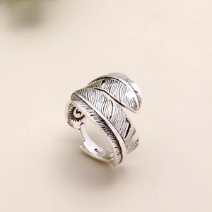 May include: A silver ring featuring a feather design. The ring wraps around the finger with two overlapping feather shapes. The feathers have detailed lines and textures. The ring is set against a neutral background.