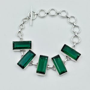 Può includere: Un bracciale color argento con cinque pietre rettangolari sfaccettate color verde smeraldo incastonate in cornici di metallo. Il bracciale è composto da una serie di maglie circolari e si chiude con una chiusura a barra a T.