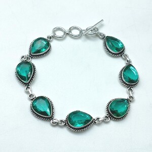 Op de afbeelding: Een zilverkleurige armband met zes peervormige, gefacetteerde groene stenen in gedraaide metalen zettingen. De schakels zijn verbonden door kleine ronde ringetjes en het sieraad wordt gesloten met een staaf- en ringvormige knevelsluiting.