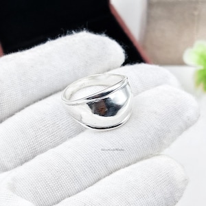 May include: A polished silver ring with a wide, smooth, and slightly domed band. The metal surface is highly reflective, showing subtle highlights and shadows. The text SilverGoldWorks is printed in small black letters on the white fabric surface.