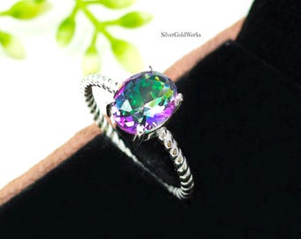 May include: A silver ring with a large, oval-cut, iridescent gemstone. The gemstone displays a spectrum of colors, including green, purple, and blue. The ring's band has a twisted rope design. The background is black and brown.