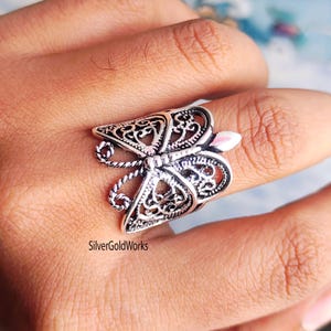 Puede incluir: Un anillo de mariposa plateado con intrincados detalles de filigrana. El anillo presenta alas caladas y un dise&ntilde;o delicado, con un toque de rosa en las antenas. El anillo se lleva en un dedo.