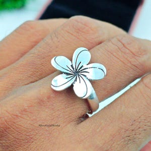 Può includere: Un anello color argento con un design a fiore. Il fiore ha cinque petali bordati di nero, con dettagli neri al centro. L'anello è indossato su un dito.