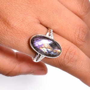 Op de afbeelding: Een zilveren ring met een ovale, iriserende edelsteen met paarse, gouden en blauwe tinten. De ring heeft een decoratieve band met kleine, parelachtige details. De edelsteen is ongeveer 2,5 cm lang.