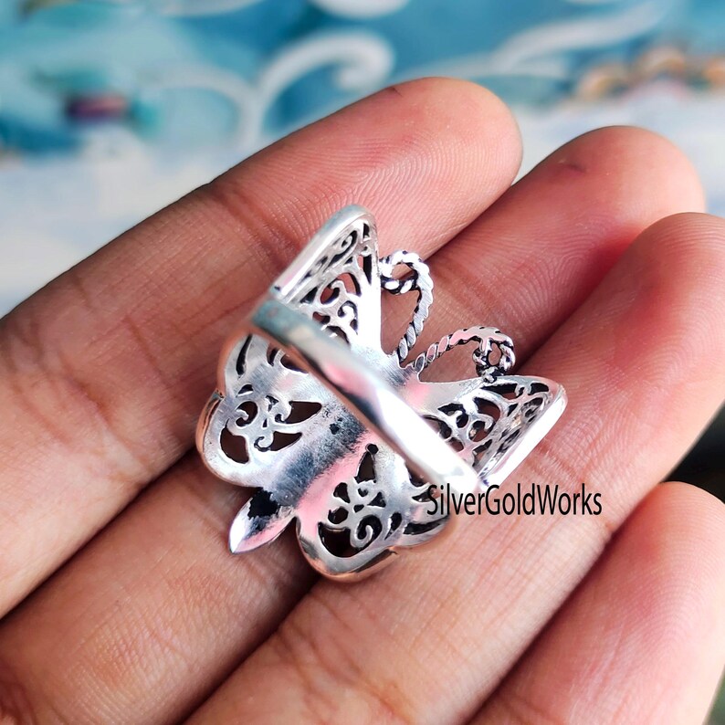 Puede incluir: Un anillo de plata con forma de mariposa y un intrincado dise&ntilde;o calado. El anillo presenta una forma de mariposa detallada con delicados patrones recortados. El anillo se muestra desde la parte posterior, revelando la banda y la parte inferior de la mariposa.