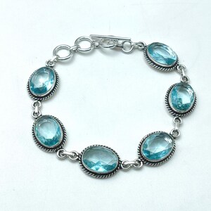 Op de afbeelding: Een zilveren schakelarmband met vijf ovale, gefacetteerde lichtblauwe edelstenen. Elke steen is gezet in een gedraaide zilveren rand en verbonden door ronde zilveren schakels, afgesloten met een zilveren knevelsluiting aan de bovenzijde.