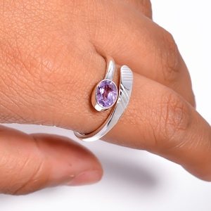 Może przedstawiać: Srebrny pierścionek z owalnym, jasnofioletowym kamieniem szlachetnym. Pierścionek ma unikalny, otwarty design z zakrzywionym paskiem, który owija się wokół palca. Kamień szlachetny jest osadzony w oprawie.