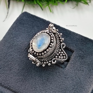 Puede incluir: Un anillo de piedra lunar ovalada con una piedra cabujón pulida e iridiscente engastada en un marco de plata oxidada detallado con texturas metálicas de cuentas y cuerdas. El anillo incluye un diseño de relicario y el texto SilverGoldWorks.