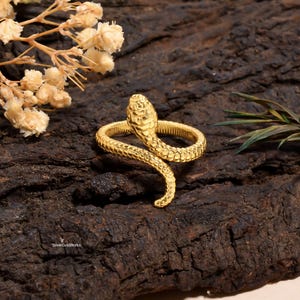 Puede incluir: Un anillo de serpiente dorado con escamas detalladas y un diseño en espiral. La cabeza de la serpiente está levantada, sobre un fondo oscuro y texturizado. El anillo es una joya.