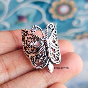 Puede incluir: Un anillo plateado con forma de mariposa con intrincados detalles de filigrana. El anillo presenta un dise&ntilde;o de mariposa detallado con alas caladas y un cuerpo texturizado. El anillo se sostiene en una mano sobre un fondo borroso.
