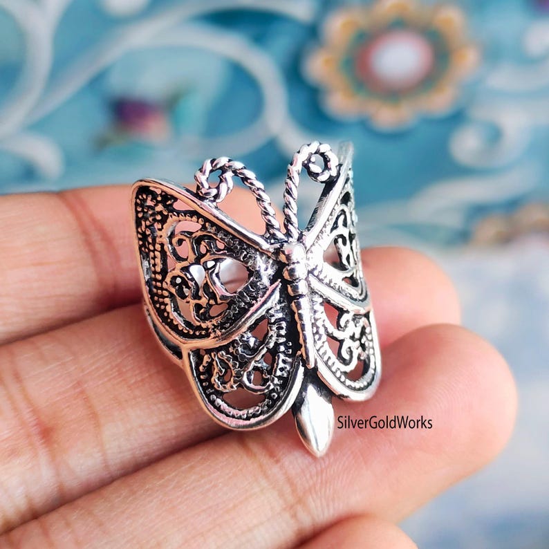 Puede incluir: Un anillo plateado con forma de mariposa con intrincados detalles de filigrana. El anillo presenta un dise&ntilde;o de mariposa detallado con alas caladas y un cuerpo texturizado. El anillo se sostiene en una mano sobre un fondo borroso.