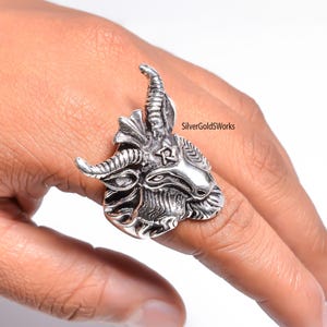 May include: A silver-colored ring featuring a detailed goat head design. The ring has prominent horns, a beard, and intricate facial features. The ring is worn on a finger, showcasing the craftsmanship and style. The text "SilverGoldWorks" is visible.