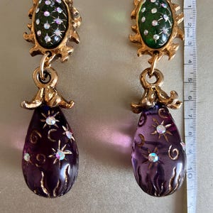 Könnte beinhalten: Ein Paar goldfarbene Vintage-Ohrringe. Jeder Ohrring hat ein grünes ovales Oberteil mit weißen Akzenten, das mit einem tropfenförmigen lila Anhänger verbunden ist. Die Anhänger sind mit sternförmigen und wirbelförmigen Details versehen. Die Ohrringe sind ca. 7.6 cm lang.