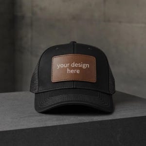 May include: Black trucker hat with a brown leather patch. The patch features the text "your design here" in white. The hat has a mesh back and a curved brim. The hat is sitting on a gray surface.