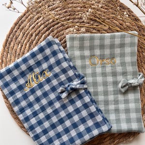 Peut inclure: Deux couvertures pour bébé à motif vichy. L'une est bleue et blanche, brodée de "Alix" en fil d'or. L'autre est grise et blanche, brodée de "Orso" en or. Chaque couverture a un petit nœud.