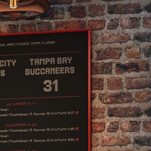 Op de afbeelding: Een scorebord met de score van 31 voor de Tampa Bay Buccaneers. De tekst bevat "Tampa Bay Buccaneers" en wedstrijddetails. Het bord is rood omlijst en staat tegen een bakstenen muur.
