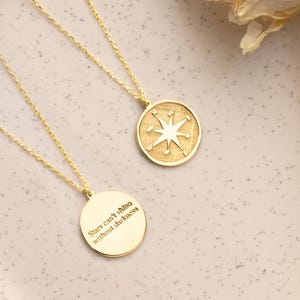 May include: Two gold necklaces with pendant charms on a light pink plate. One pendant has a compass design, the other has the engraved text "Stars can't shine without darkness". The necklaces are displayed with dried flowers and a white cloth.