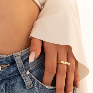 May include: A gold ring with the word "Love" inscribed. The rectangular ring is worn on a finger. The person is wearing blue denim jeans and a white blouse.