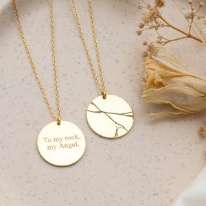 May include: Two gold-colored pendant necklaces. One pendant has the engraved text "To my rock, my Angel." The other pendant has a cracked design. The necklaces are on a light pink plate with dried flowers.