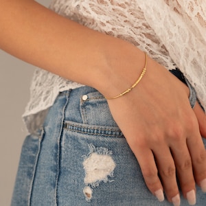May include: A fine gold bracelet featuring small beads and cylindrical details. The bracelet is worn on a wrist, set against a white lace top and blue denim jeans.