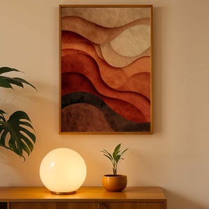 May include: A framed abstract art piece with layered, wavy shapes in shades of red, brown, and beige hangs on a cream-colored wall. A round, glowing lamp sits on a wooden cabinet, with a potted plant to the left and a small plant in an orange pot to the right.