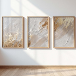 May include: Three framed abstract artworks. Each piece displays a textured design with white, beige, and gold hues. The art is set within light brown wooden frames, forming a coordinated set for wall decoration.