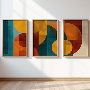 Peut inclure: Trois impressions d'art abstraites encadrées. Chaque impression présente un design géométrique avec des formes superposées en bleu sarcelle, orange, jaune et bordeaux. Les cadres sont en bois clair et l'œuvre est exposée sur un mur blanc.